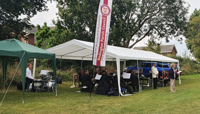 Photo depicts people playing brass instruments under a white gazebo placed on green grass.