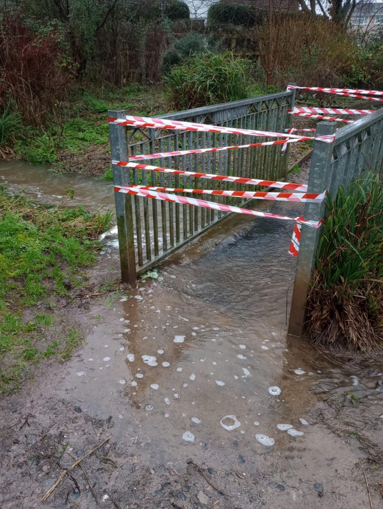 A footbridge taped off with red and white hazard tape, stream water blocks the pathway.