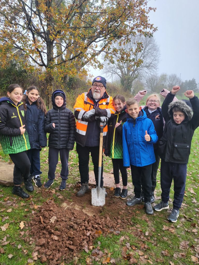 Member of Council staff holds a spade, several school children are stood with him.  They are in a park.