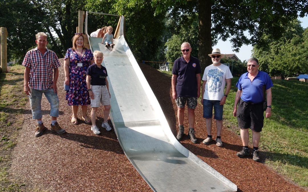 Eight people by a slide, two of them are at the top of the slide. There are trees behind and a blue sky.