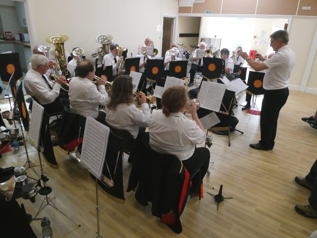 A group of musicians in uniform play a variety of brass instruments in a hall, they are sat down on chairs and they are being led by a conductor who is stood before them.